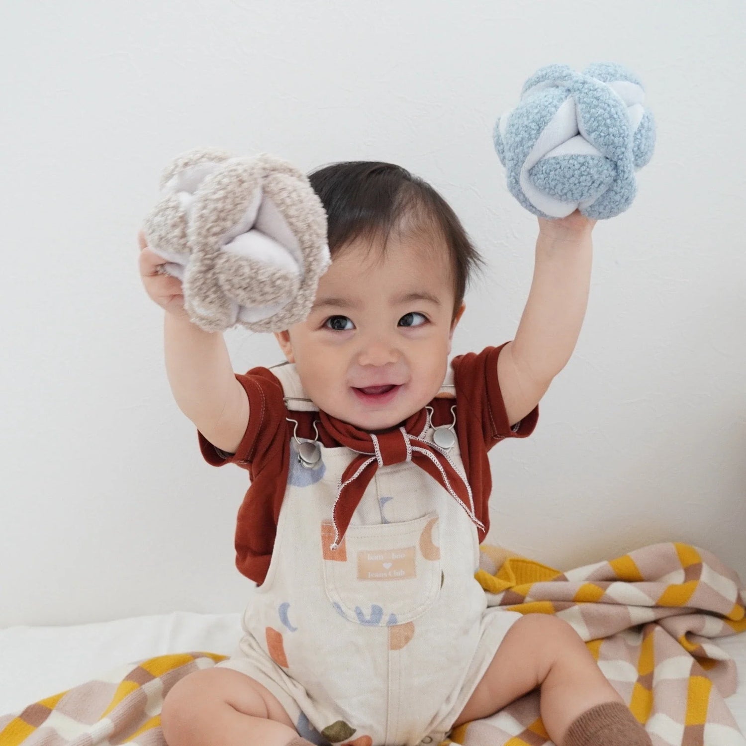 Smiling baby holding two Monti soft fabric balls, wearing a brown top and patterned overalls, sitting on a checkered blanket.