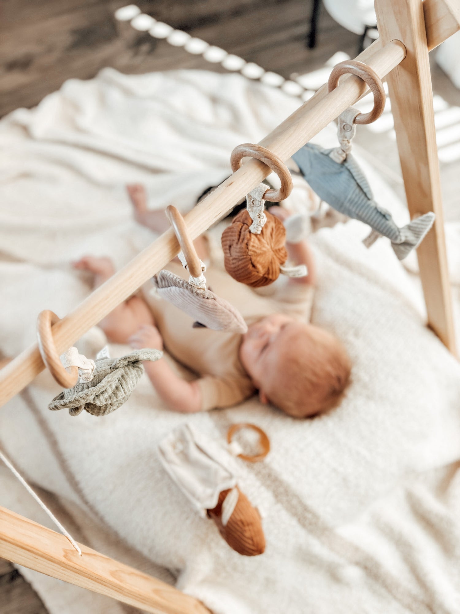 Baby lying on a cozy blanket under a wooden play gym with Little Lamb Kind toy rattles, featuring soft, textured food and animal shapes.